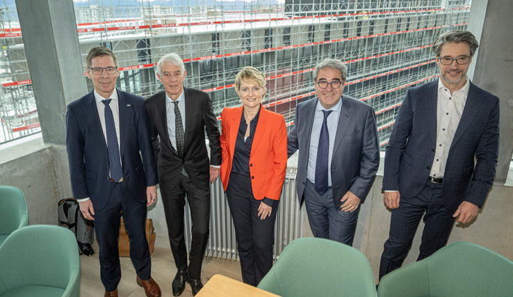 Les personnages posent dans une salle de conférence très vitrée qui donne sur le bâtiment en construction.