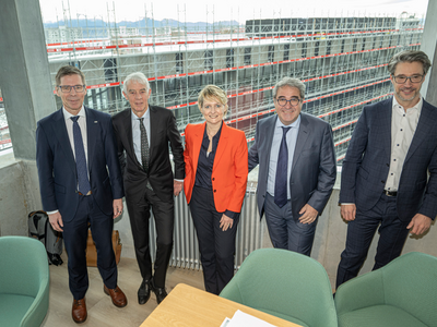 Les personnages posent dans une salle de conférence très vitrée qui donne sur le bâtiment en construction.