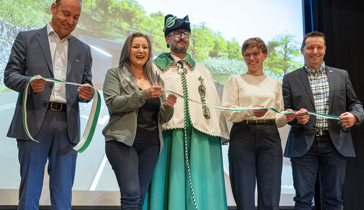 Les personnages posent sur une scène devant un écran qui montre le futur tunnel qui sera construit au-dessous de Sainte-Croix