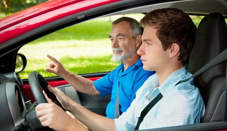 Un jeune conducteur au premier plan, qui regarde la route; sur le siège passage, un moniteur d'auto-école, l'index dressé.