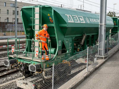 Un homme en combinaison orange sur le marchepied d'un wagon de granulats de la compagnie MCB