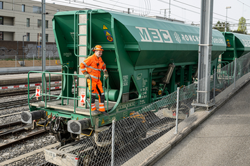 Un homme en combinaison orange sur le marchepied d'un wagon de granulats de la compagnie MCB