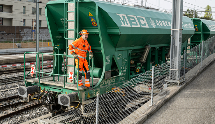 Un homme en combinaison orange sur le marchepied d'un wagon de granulats de la compagnie MCB