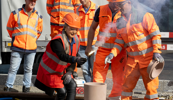 Coiffée d'un casque de chantier et vêtue d'une tenue orange comme les collaborateurs de l'entreprise de travaux publics qui l'entourent, la conseillère d'Etat tient une mèche enflammée qui va embraser l'appareil destiné à souder les rails.