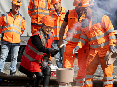 Coiffée d'un casque de chantier et vêtue d'une tenue orange comme les collaborateurs de l'entreprise de travaux publics qui l'entourent, la conseillère d'Etat tient une mèche enflammée qui va embraser l'appareil destiné à souder les rails.