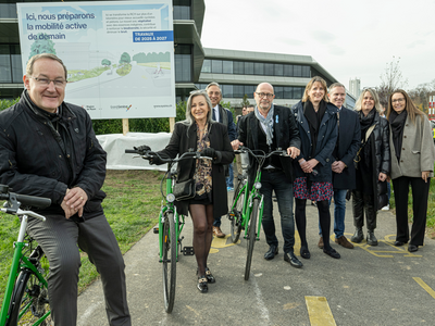 les personnes posent sur une route, appuyée sur des vélos. Derrière, au bord de la chaussée, un panneau de chantier. On lit: "Ici nous préparons la mobilité active de demain." On voit deux ouvriers casqués.