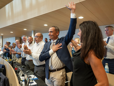 Le nouveau président à son siège, debout, applaudi par ses collègues. Il lève la main gauche et a posé la droit sur sa poitrine.