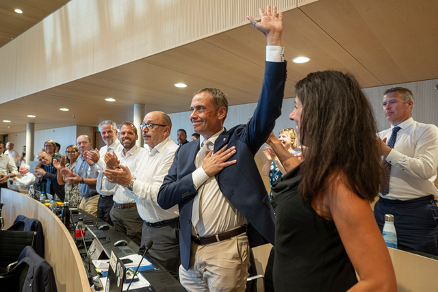 Le nouveau président à son siège, debout, applaudi par ses collègues. Il lève la main gauche et a posé la droit sur sa poitrine.