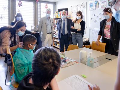 Les personnes sont debout dans une salle de classe, autour d'une table où sont assis deux enfants dont l'un porte une blouse. Tout le monde porte un masque sanitaire. 