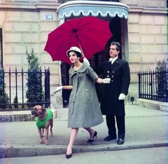 La mannequin tient un boulegogue en laisse, vêtu d'un manteau vert pour chien. Derrière elle, un homme en livrée noire tient un parapluie rouge pour abriter la mannequin.