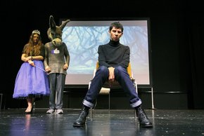 photo de spectacle: un homme assis au premier plan, qui regarde au sol. Derrière lui à gauche, une comédienne vêtue en fée, et un personnage coiffé d'une tête d'âne et dont le pull porte le logo de Facebook (pouce levé) 