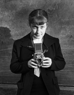 Image en noir et blanc de la photographe, qui regarde droit devant elle en tenant l'appareil à deux mains devant sa poitrine. Elle a des cheveux longs et une frange, une cravate. Derrière, un mur avec des grafitis. 