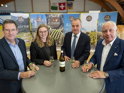 Les personnes posent autour d'une table, chacune un verre de vin blanc à la main. Au fond, des affiches publicitaires pour les différents vignobles du canton.
